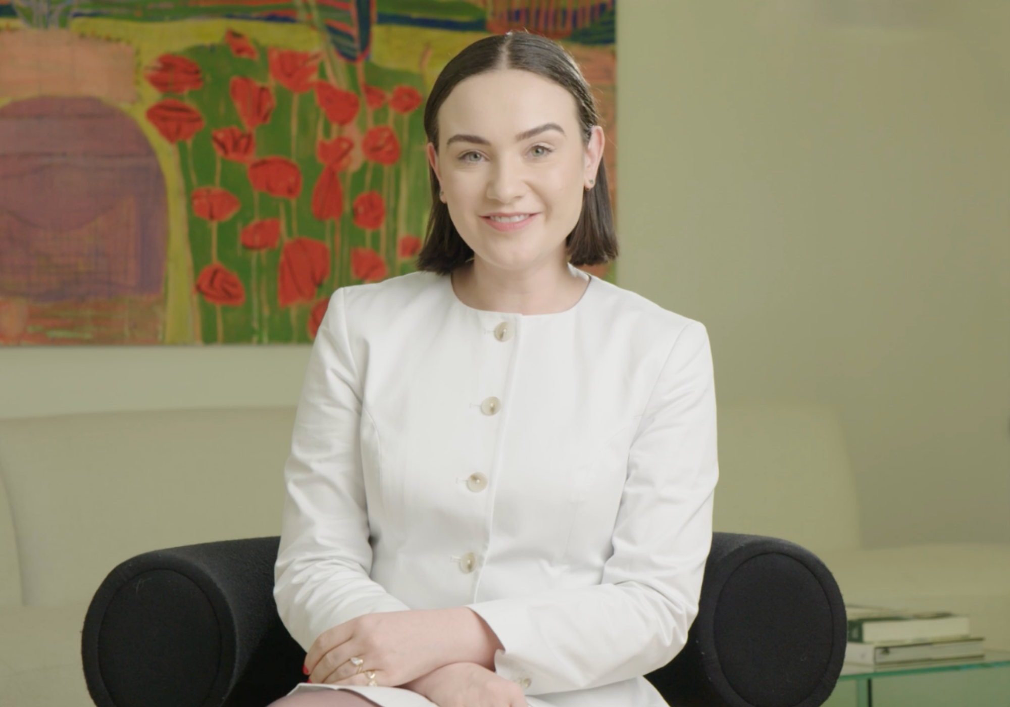 Woman in a white jacket sitting in a room with a colorful painting in the background