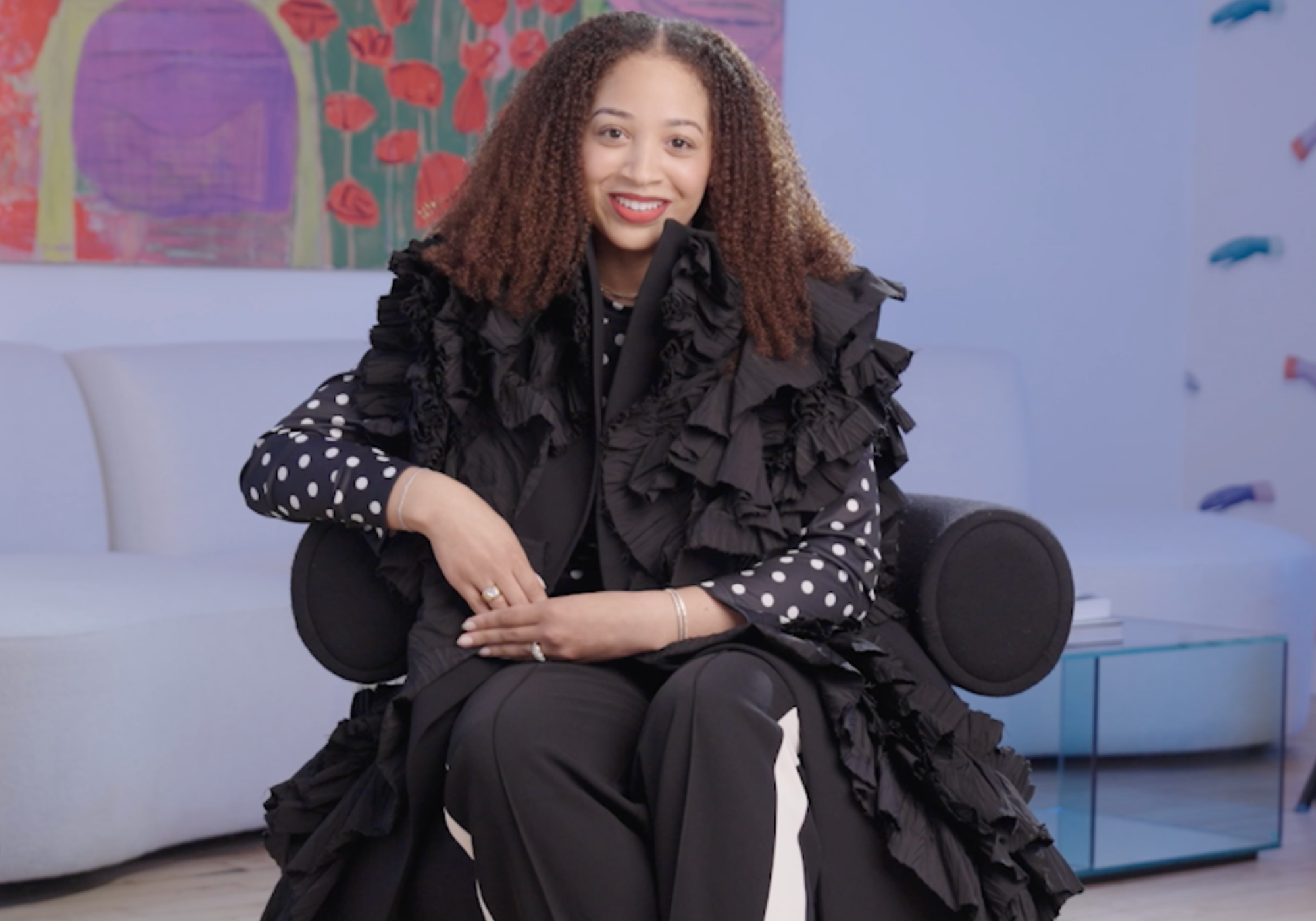 Woman in a black ruffled outfit sitting on a white chair with a colorful abstract painting in the background.