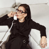 Woman in black outfit sitting on a white couch, talking on a white phone.