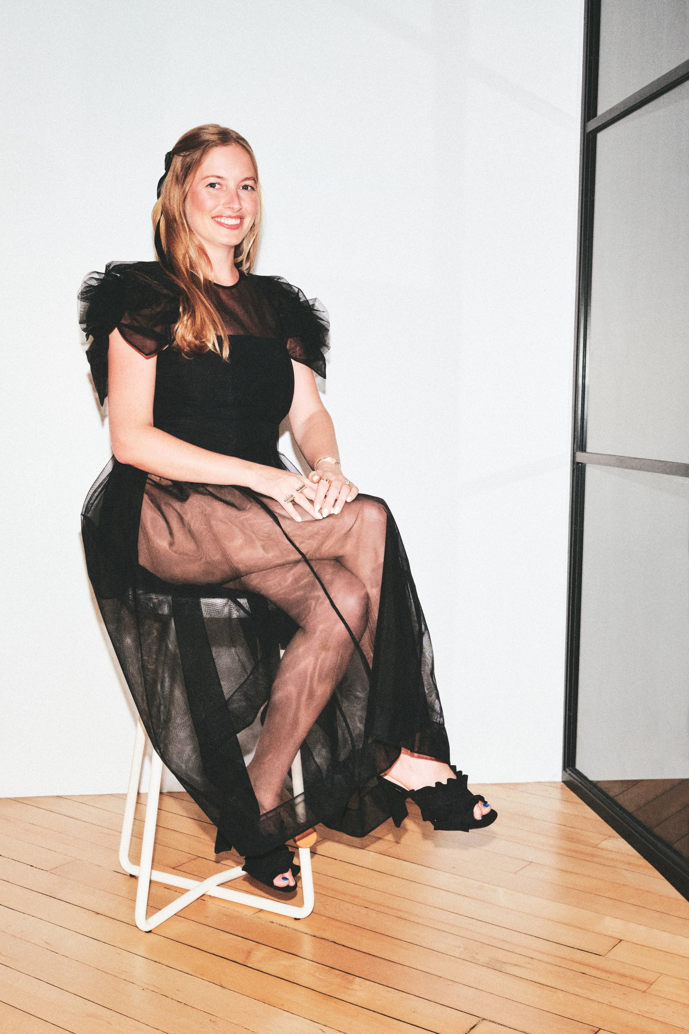 A woman sits on a white metal stool wearing a black sheer tulle dress with ruffled short sleeves and black open-toe heels, photographed indoors against a white wall with wood flooring.