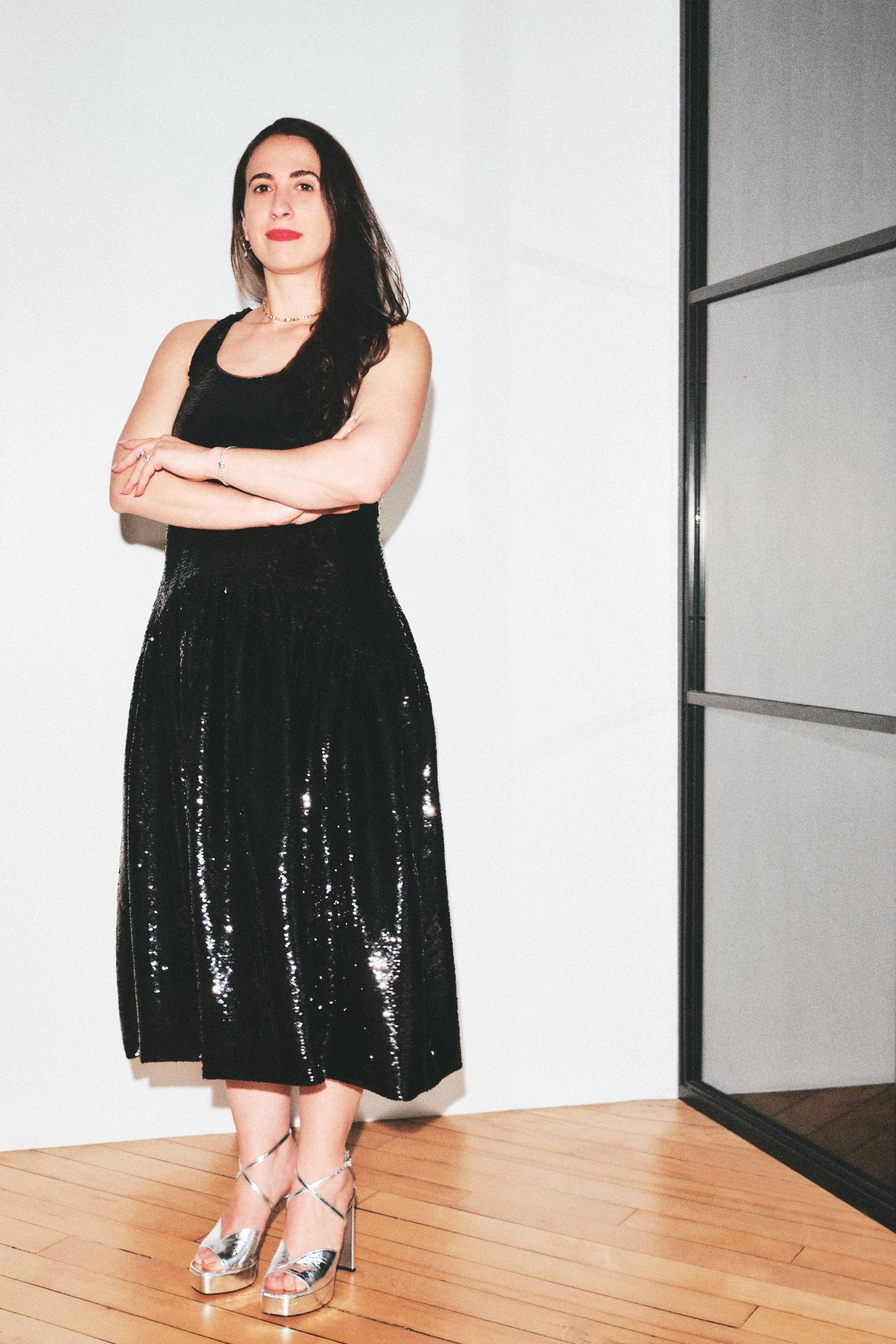 A woman stands indoors wearing a sleeveless black sequined midi dress with a scoop neckline and a flared skirt, paired with metallic high-heel sandals. She is posed with arms crossed against a white wall with a wood floor and a glass-paneled door frame to the right.