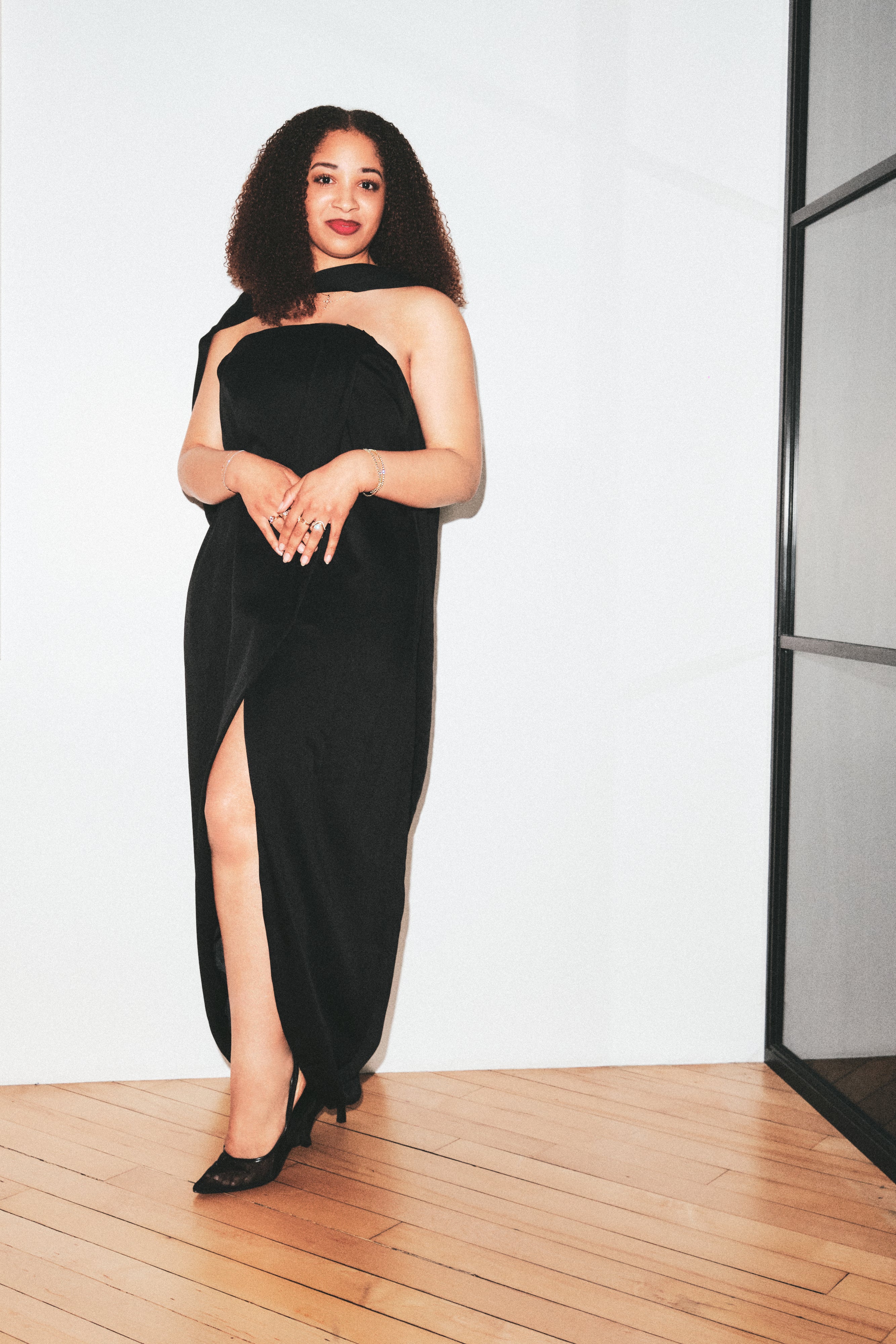 A woman wearing a black strapless velvet gown with a thigh-high slit and black pointed heels, standing on a light wood floor against a white wall.
