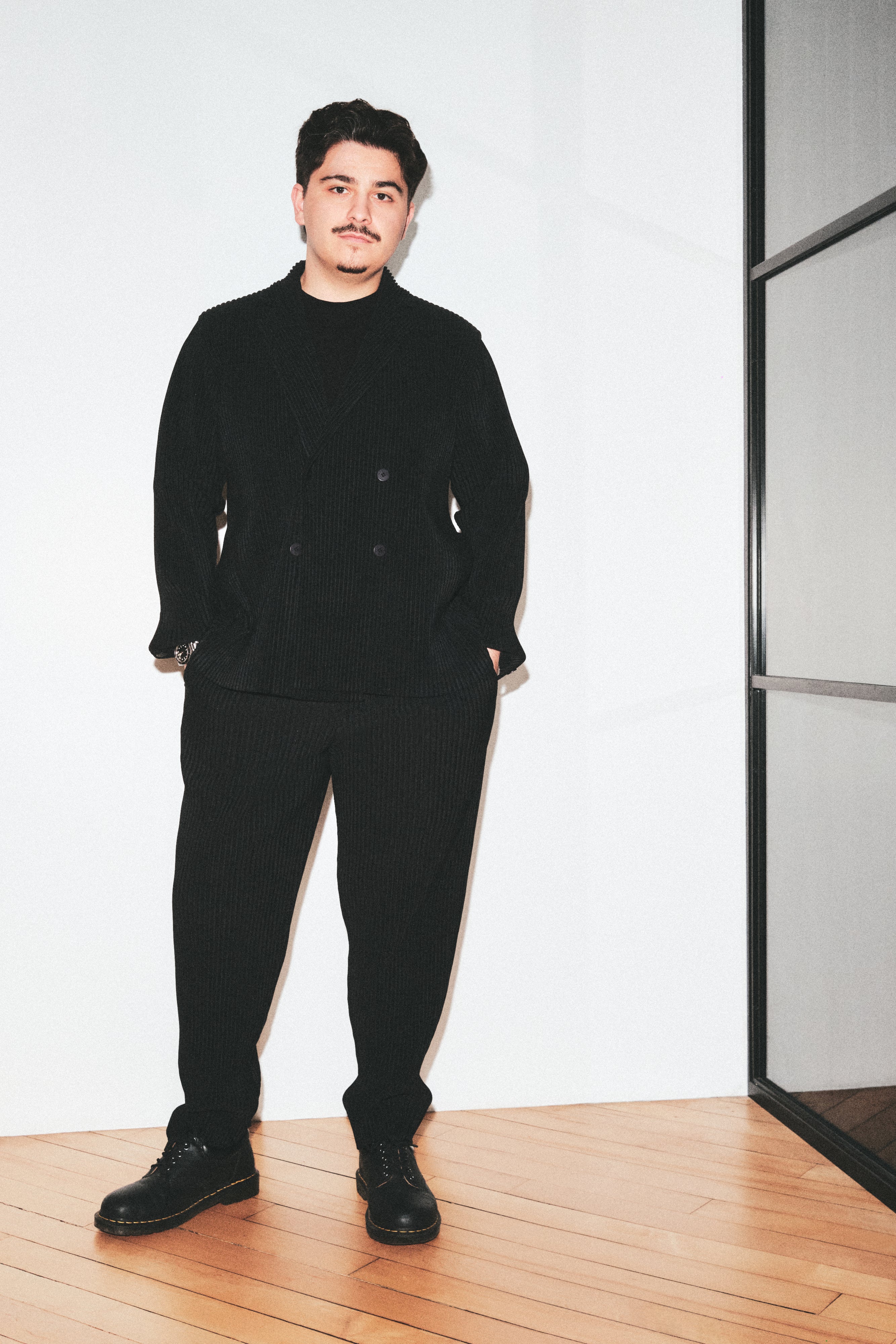 A man modeling a black suit standing on a light wood floor against a white wall.