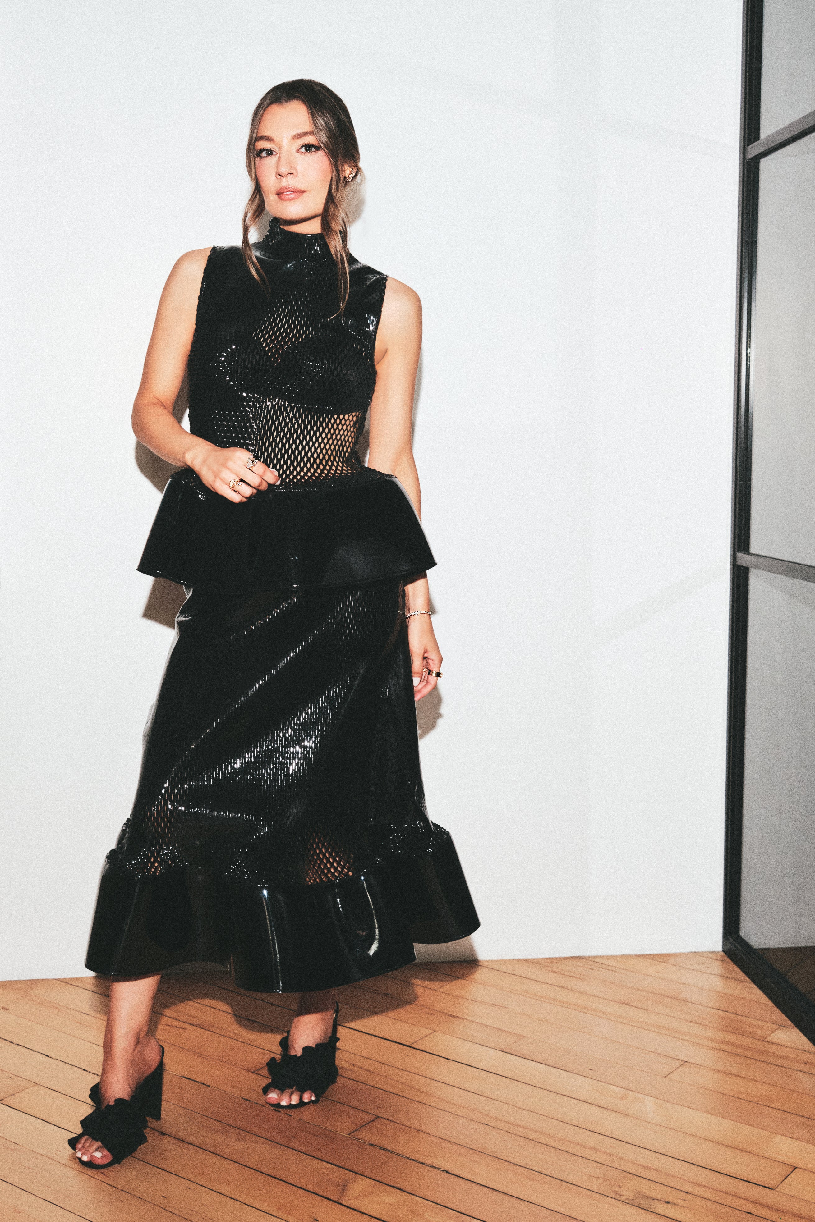 A woman models a black sleeveless midi dress with a high-neck mesh overlay, structured peplum waist, and flared ruffled hem, paired with black strappy heels, set against a minimalist white wall and light wood floor.