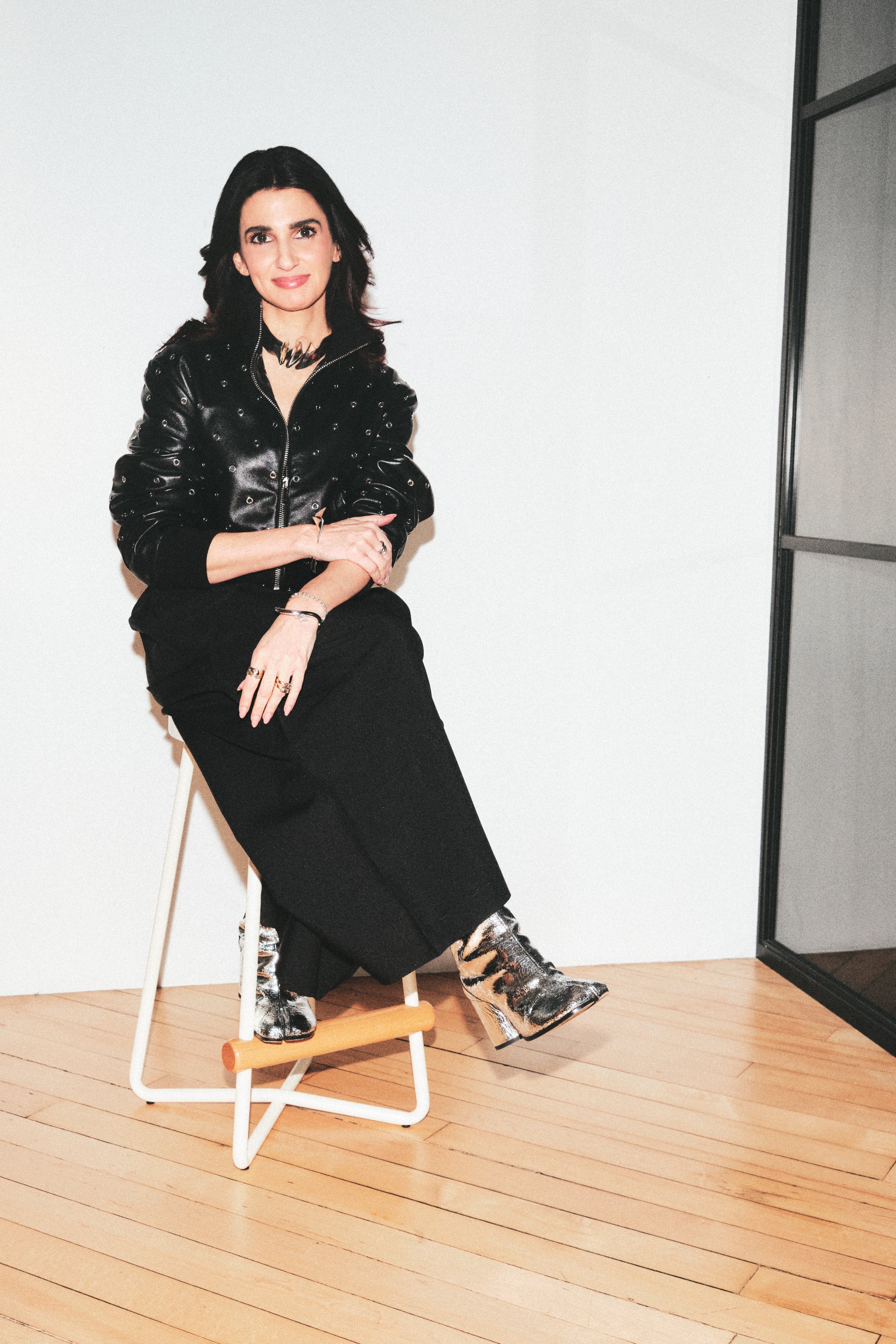 A woman sits on a white and light wood stool in a minimalist room with light wood floors and a white wall. She is wearing a black faux-fur jacket, black wide-leg pants, and black-and-silver patterned ankle boots.