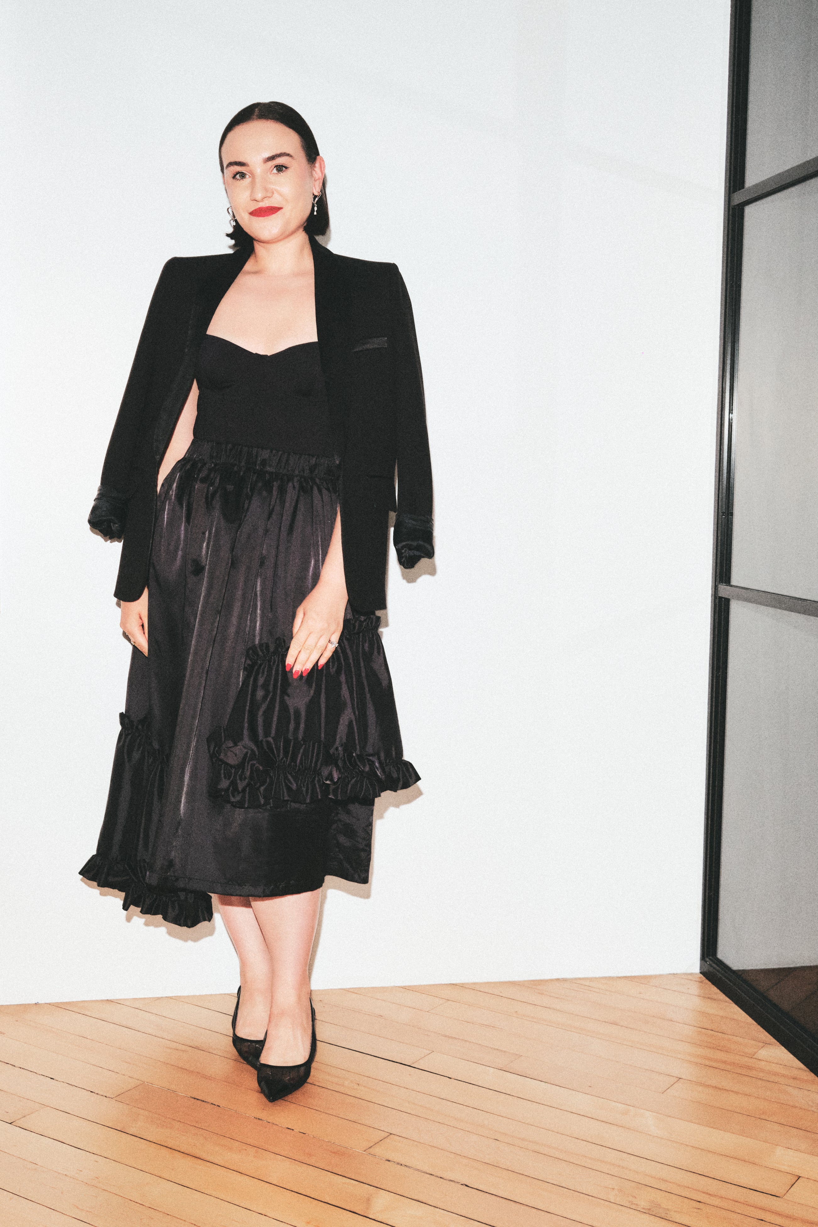 A woman modeling a black outfit consisting of a strapless black top, a black ruffled midi skirt, and a black velvet blazer, standing on a light wood floor against a white wall.