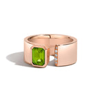 A rose gold ring with a wide band featuring a large emerald-cut green gemstone on one side and a row of small clear stones on the other, shown on a white background.