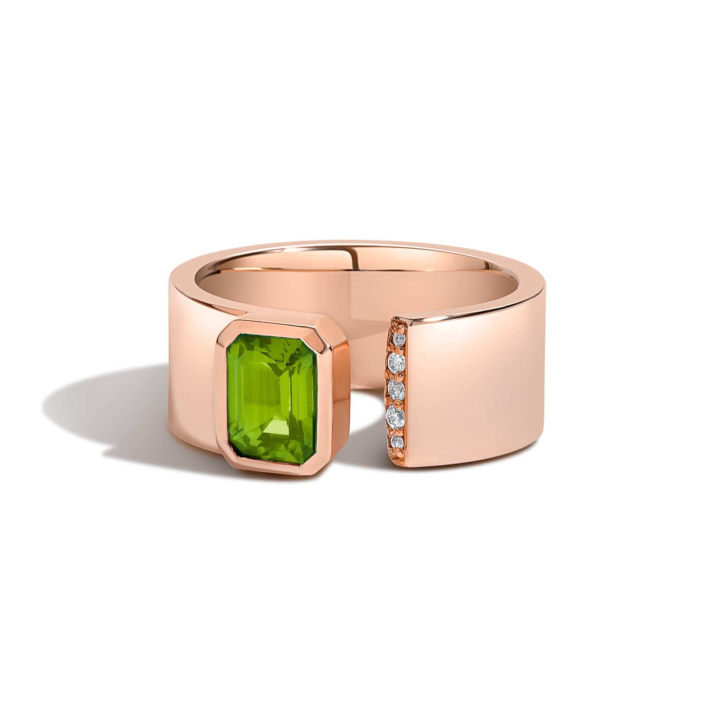 A rose gold ring with a wide band featuring a large emerald-cut green gemstone on one side and a row of small clear stones on the other, shown on a white background.