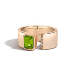 A gold ring with a wide band featuring a large rectangular green peridot gemstone on one side and a row of small clear stones on the other, shown on a white background.