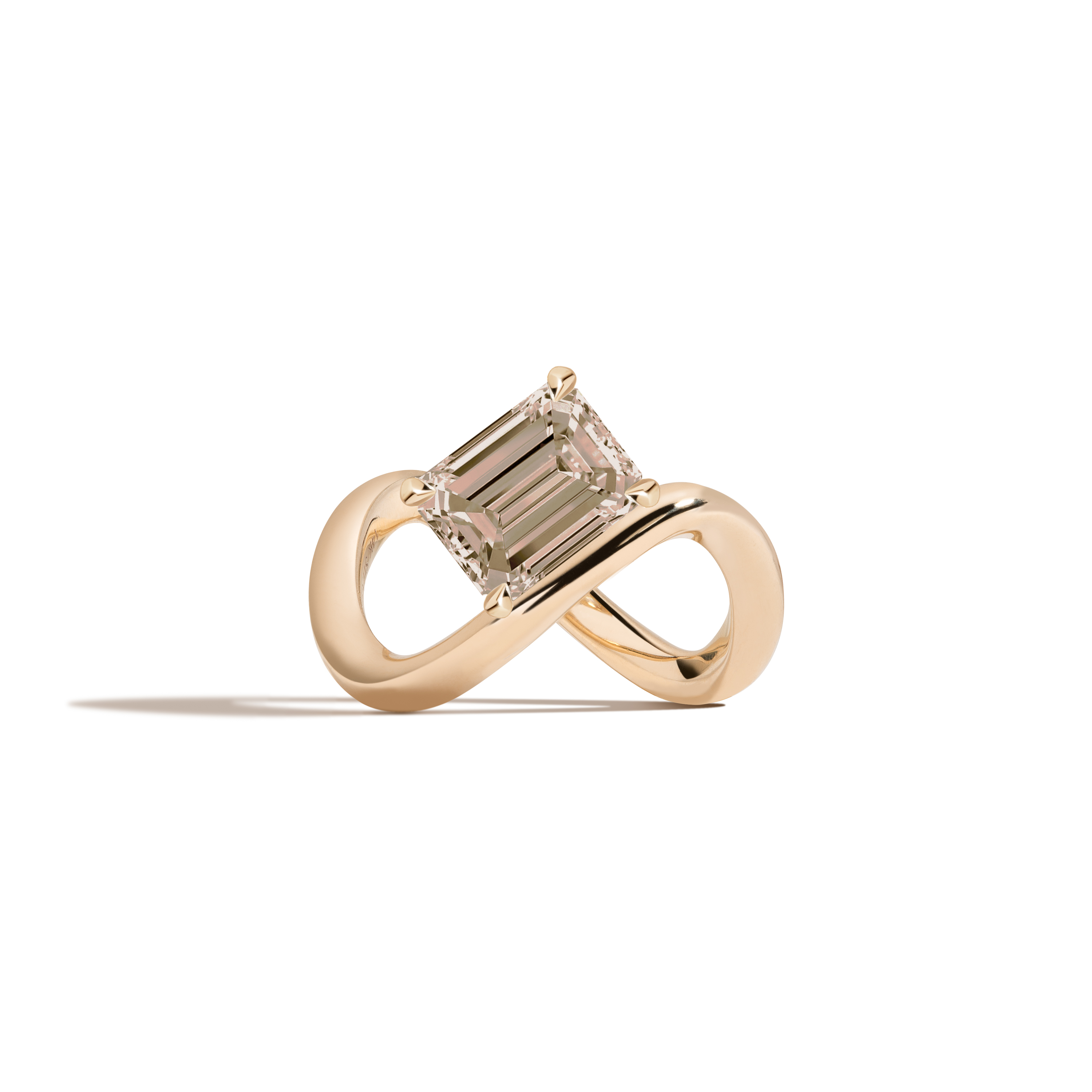A yellow gold infinity-shaped ring featuring a tilted emerald-cut champagne diamond center stone, set against a clean white background with a soft shadow.