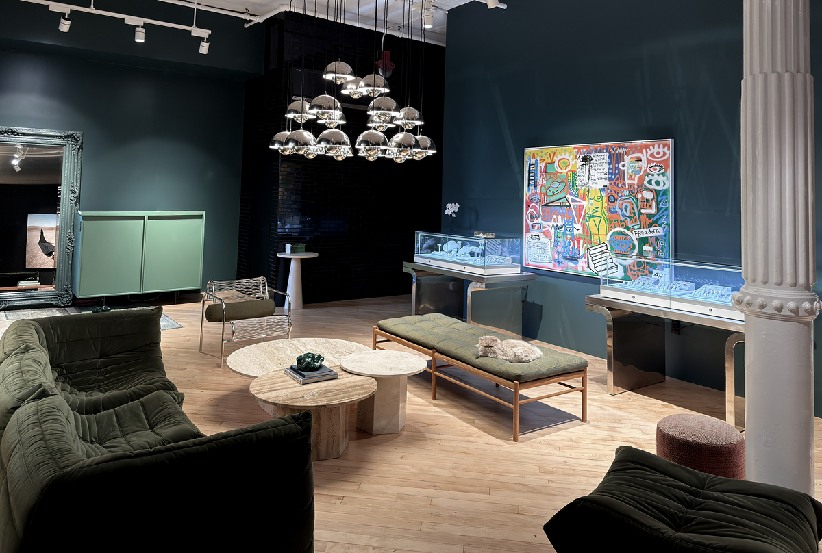 A luxury interior design showroom featuring a dark green velvet sofa, round wooden coffee tables, a green upholstered bench, and a large colorful abstract art print on a dark teal wall.