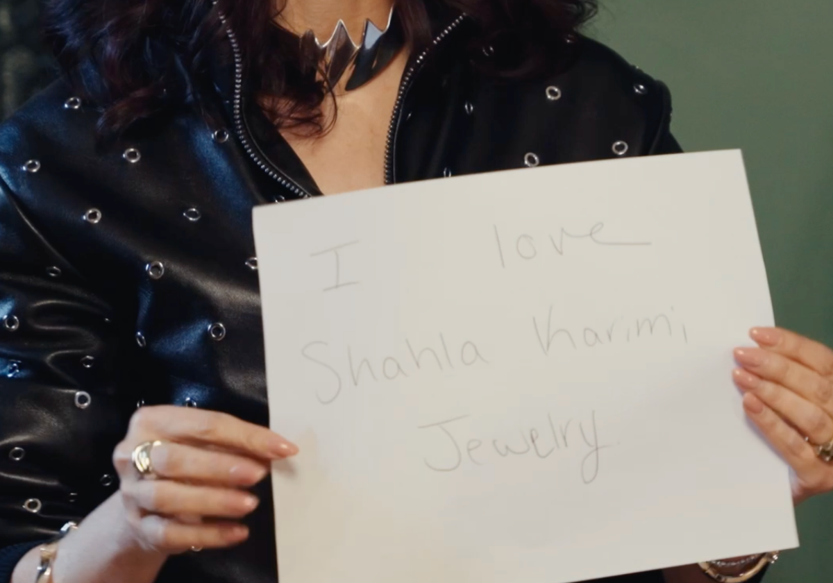 A person in a black studded leather jacket holds up a white sign that reads 'I love Shahla Karimi Jewelry' against a dark green background.