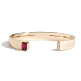 A gold cuff bracelet featuring an emerald cut ruby on one side and a series of pave-set diamonds on the other, with a glossy finish.